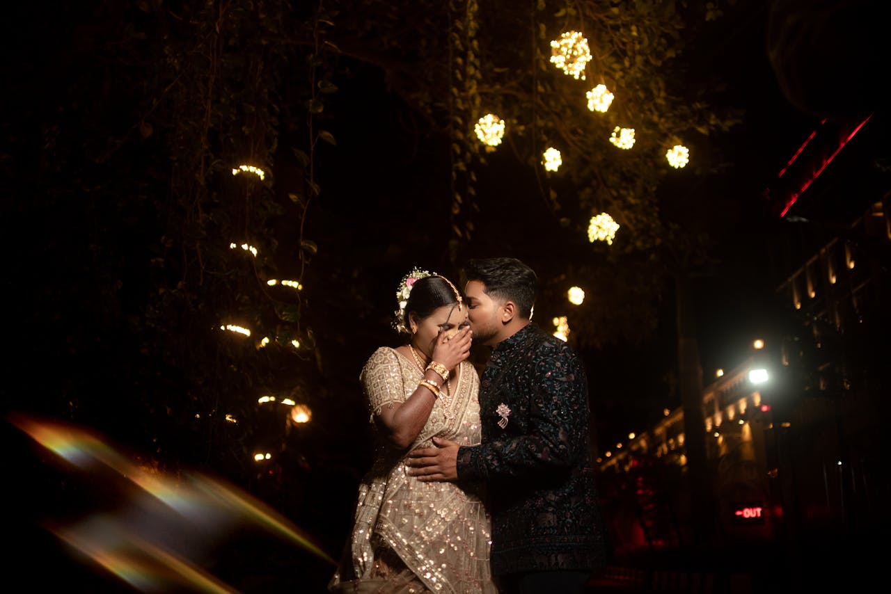 A couple shares a tender moment during a nighttime Indian wedding celebration adorned with fairy lights.